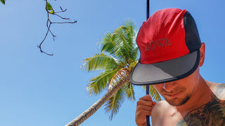 Man wearing a red and black AQWA trucker hat under a palm tree with a tropical blue sky.