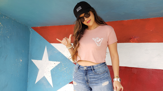 Woman wearing AQWA pink crop top and black cap, posing in front of the Puerto Rican flag.