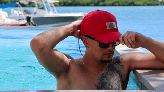 Man wearing a red AQWA cap and sunglasses while relaxing in a pool with ocean view.
