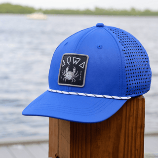AQWA royal blue mesh hat with crab patch logo, perched on a dock by the sea.
