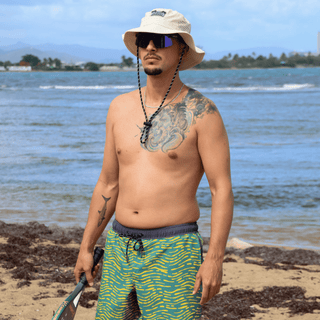 AQWA beige boonie hat and green patterned boardshorts worn by model enjoying the shore.
