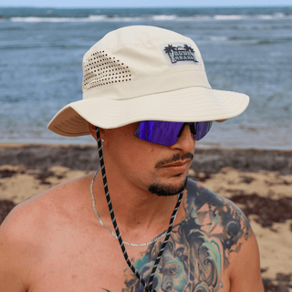 AQWA beige boonie hat with chin strap shown on model by the ocean, paired with reflective shades.