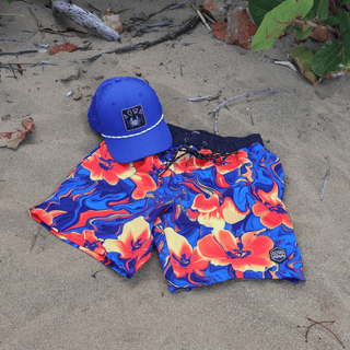 AQWA vibrant floral swim trunks and royal blue mesh cap laid on sand, inspired by island vibes and bold beach style.