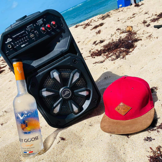 AQWA red flat-brim cap beside speaker and vodka bottle on sandy beach — summer essentials for party vibes and coastal lifestyle.
