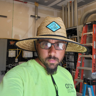 AQWA straw lifeguard hat with blue wave patch logo, worn indoors by man on worksite — showcasing sun protection and coastal-inspired design.