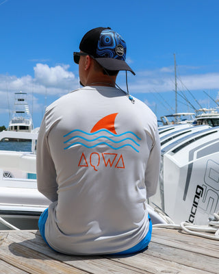 Man sited on dock in front of a boat during a sunny day, wearing a fishing AQWA rash guard and hat 